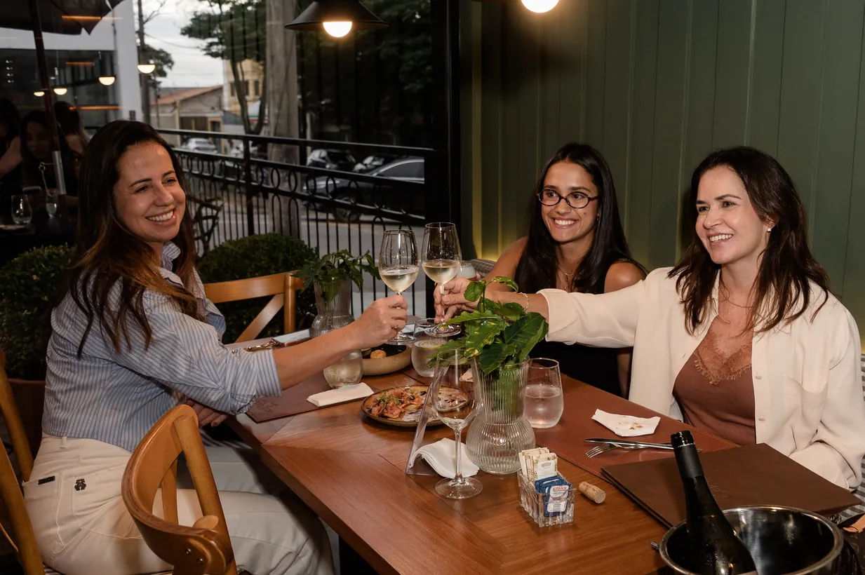Três amigas brindando à mesa, com a parede verde da Casa ao fundo
